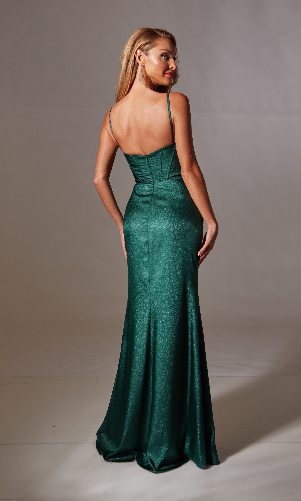 A woman faces away in a fitted, floor-length dark green Cowl-Neck Long Glitter Prom Dress (Aerie Couture 232) with a corset bodice and thin straps, posed against a plain studio backdrop.